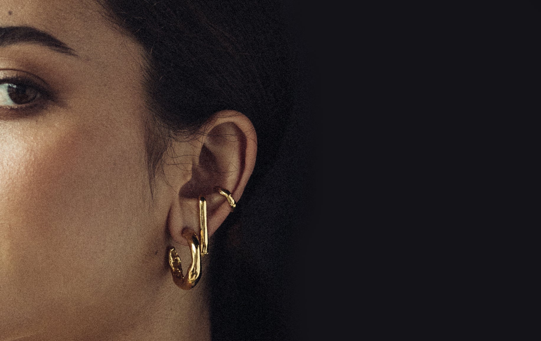 Close-up of a model wearing layered gold earrings, including textured hoops and sculptural drop designs, set against a dark background.