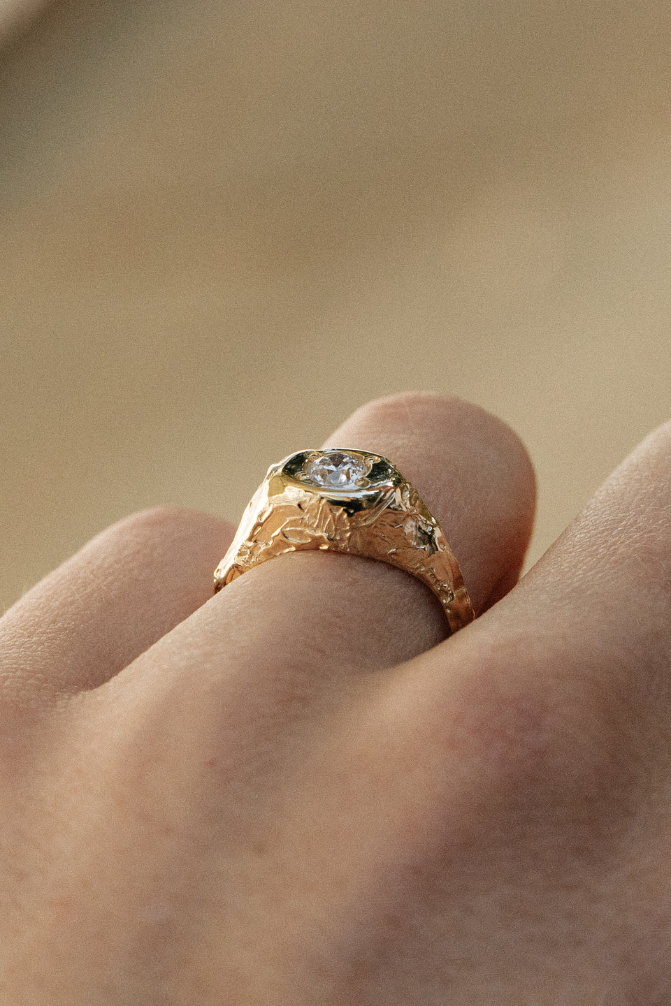 Gold signet ring with a textured, organic design and a round brilliant diamond centerpiece, displayed on a finger.